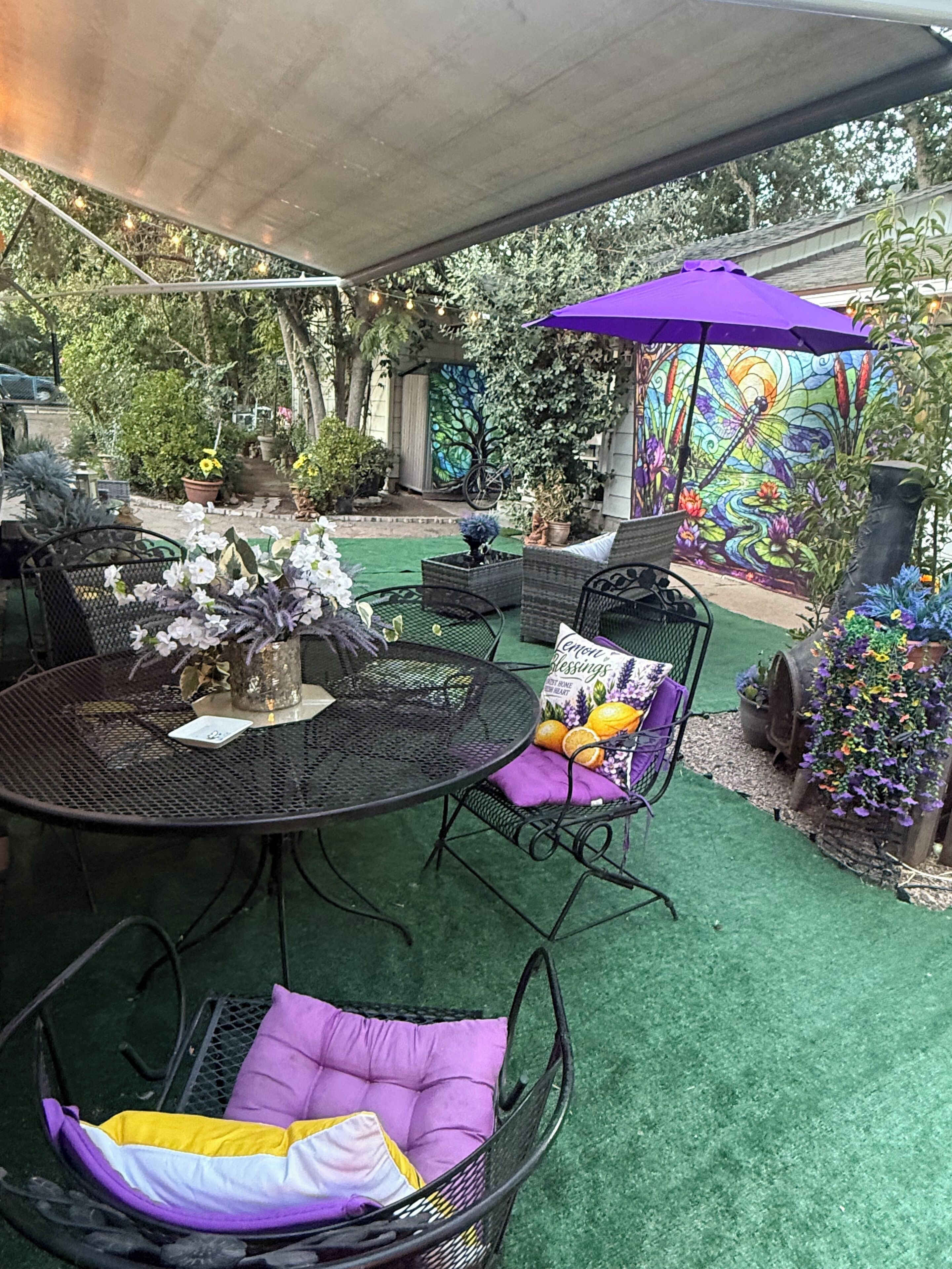 An outdoor patio area with a round table and chairs, surrounded by greenery and a decorative mural.