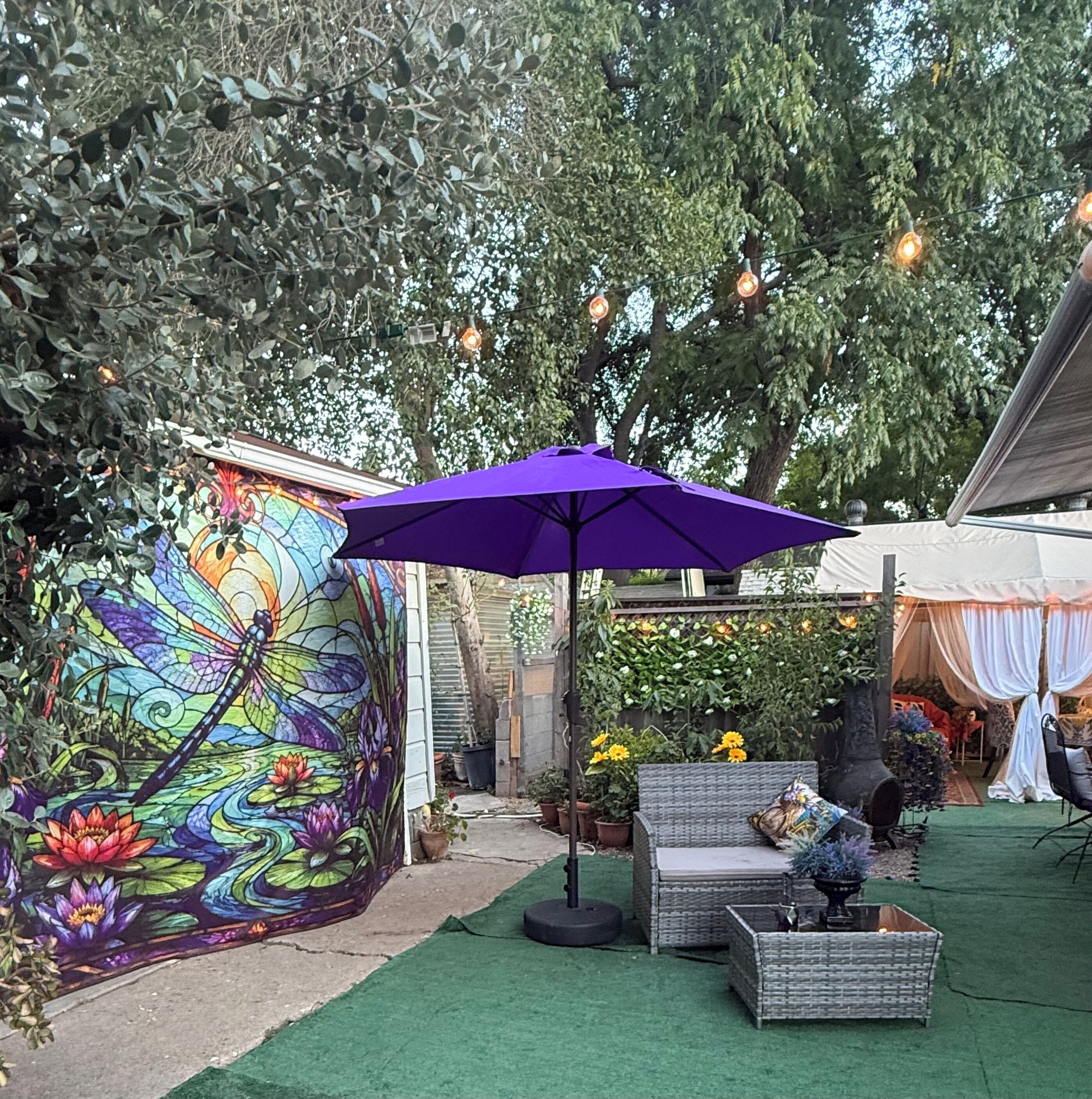 An outdoor seating area with a purple umbrella, a decorative wall mural, and a garden setting.