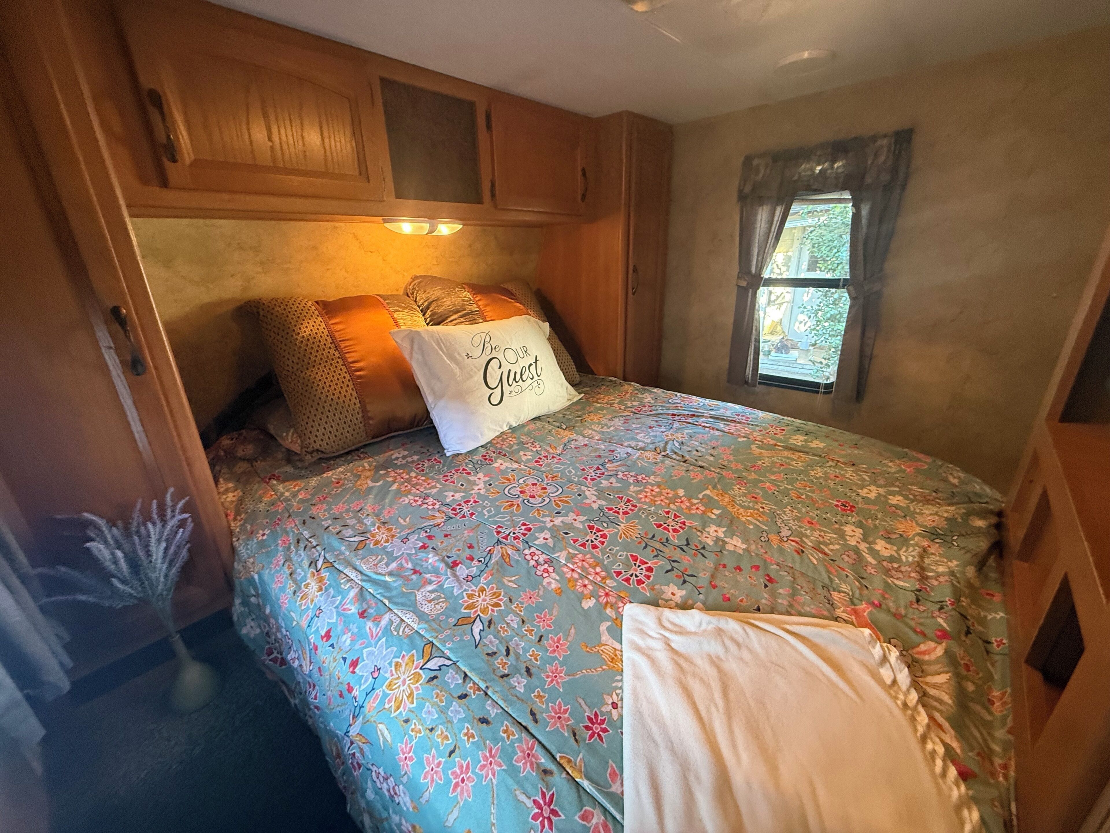 A bedroom with a floral bedspread, a pillow with 'Be Our Guest' text, wooden cabinets, and a window with a view.