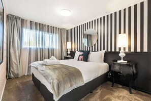 A hotel room with a large bed, a bedside table with a lamp, a mirror, and a window with curtains. - Winifreds Apartments - By Superior Stays (Hamilton)