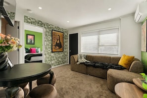 A living room with a green accent wall, a brown sofa, a dining table, and a window with blinds. - Winifreds Apartments - By Superior Stays (Hamilton)