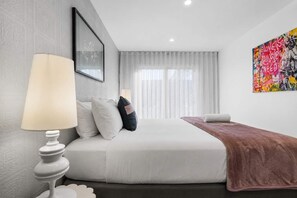 Une chambre d’hôtel moderne, équipée d’un lit, d’une table de chevet, d’une lampe, d’une fenêtre avec des rideaux et d’une œuvre d’art colorée accrochée au mur.