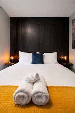 A hotel room with a bed, two bedside lamps, a headboard, and a rolled towel on the bed. - Winifreds Apartment Where Design Meets Convenience (Hamilton)