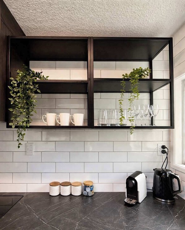 A kitchen with a black countertop, white tiled backsplash, a coffee machine, and a hanging plant. - Winifreds Apartment Where Design Meets Convenience (Hamilton)