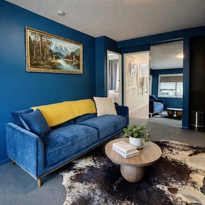 A room with a blue sofa, a round wooden coffee table with a book and a plant, a painting on the wall, and a cowhide rug. - Winifreds Apartment Where Design Meets Convenience (Hamilton)