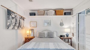 A bedroom with a bed, bedside tables, a wall-mounted shelf with baskets, and a door with shutters. - Coastal Canopy Cottage, South of 30A, Walk to Beach, Fenced Yard (Santa Rosa Beach)