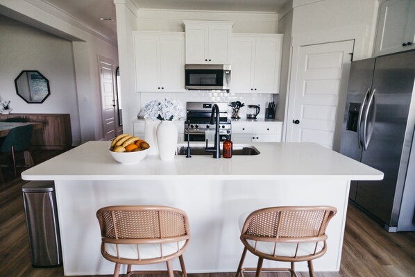 Une cuisine moderne avec des meubles blancs, un îlot central, des appareils en acier inoxydable et des chaises en osier.
