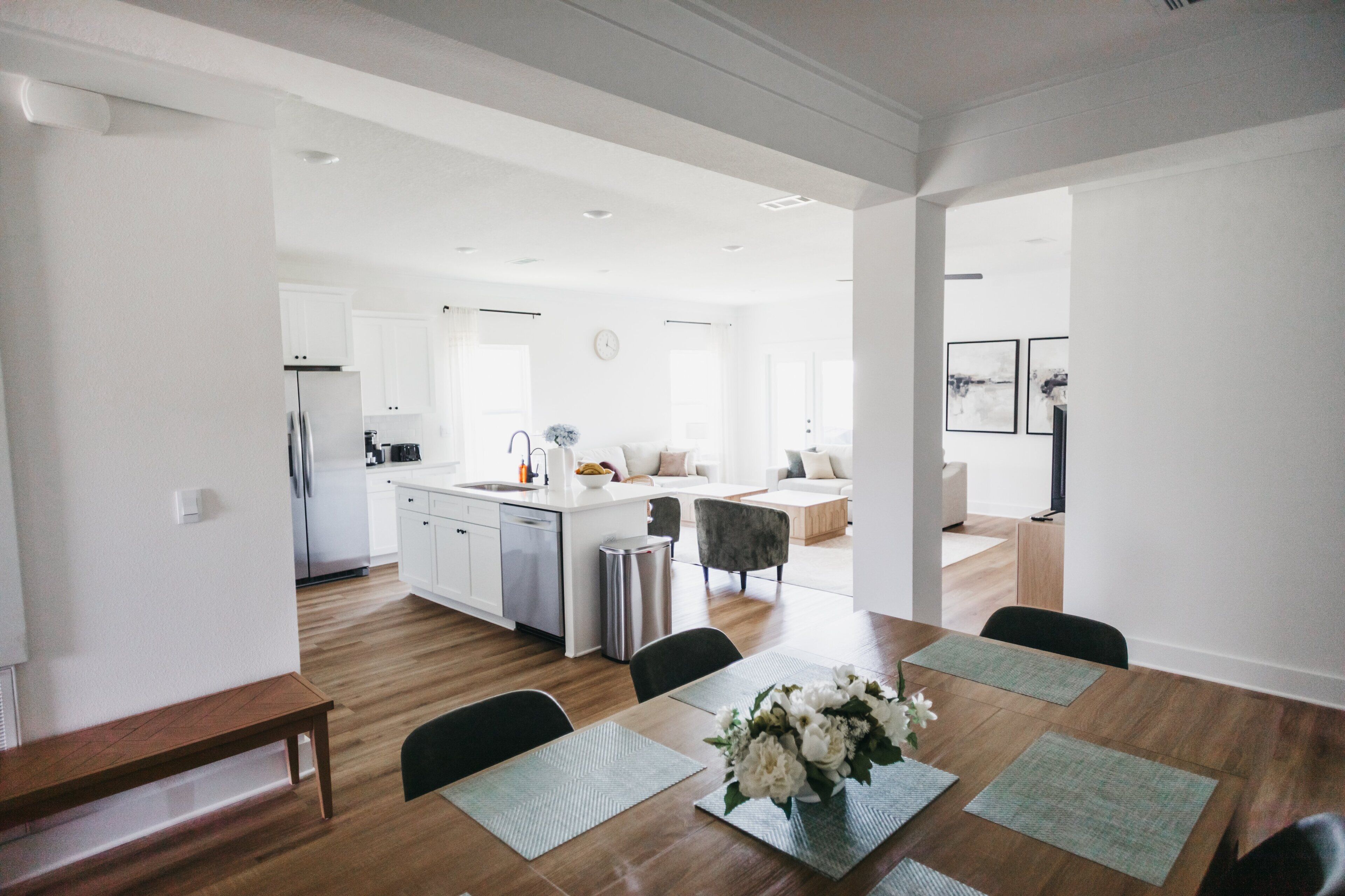 Una cocina moderna con armarios blancos, un refrigerador de acero inoxidable y un comedor con mesa y sillas de madera.