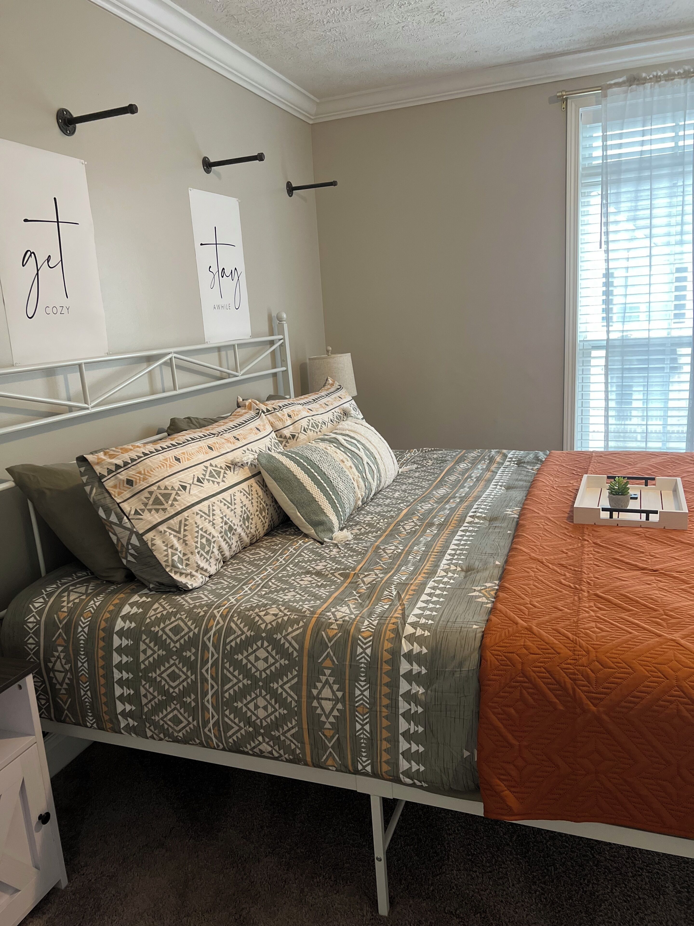 A bedroom with a bed, pillows, a bedside table, a lamp, and a window with blinds.