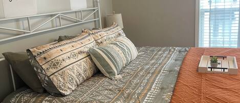 A bedroom with a bed, pillows, a bedside table, a lamp, and a window with blinds.