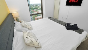 A modern hotel room with a large bed, a TV mounted on the wall, and a view of the city through a large window.