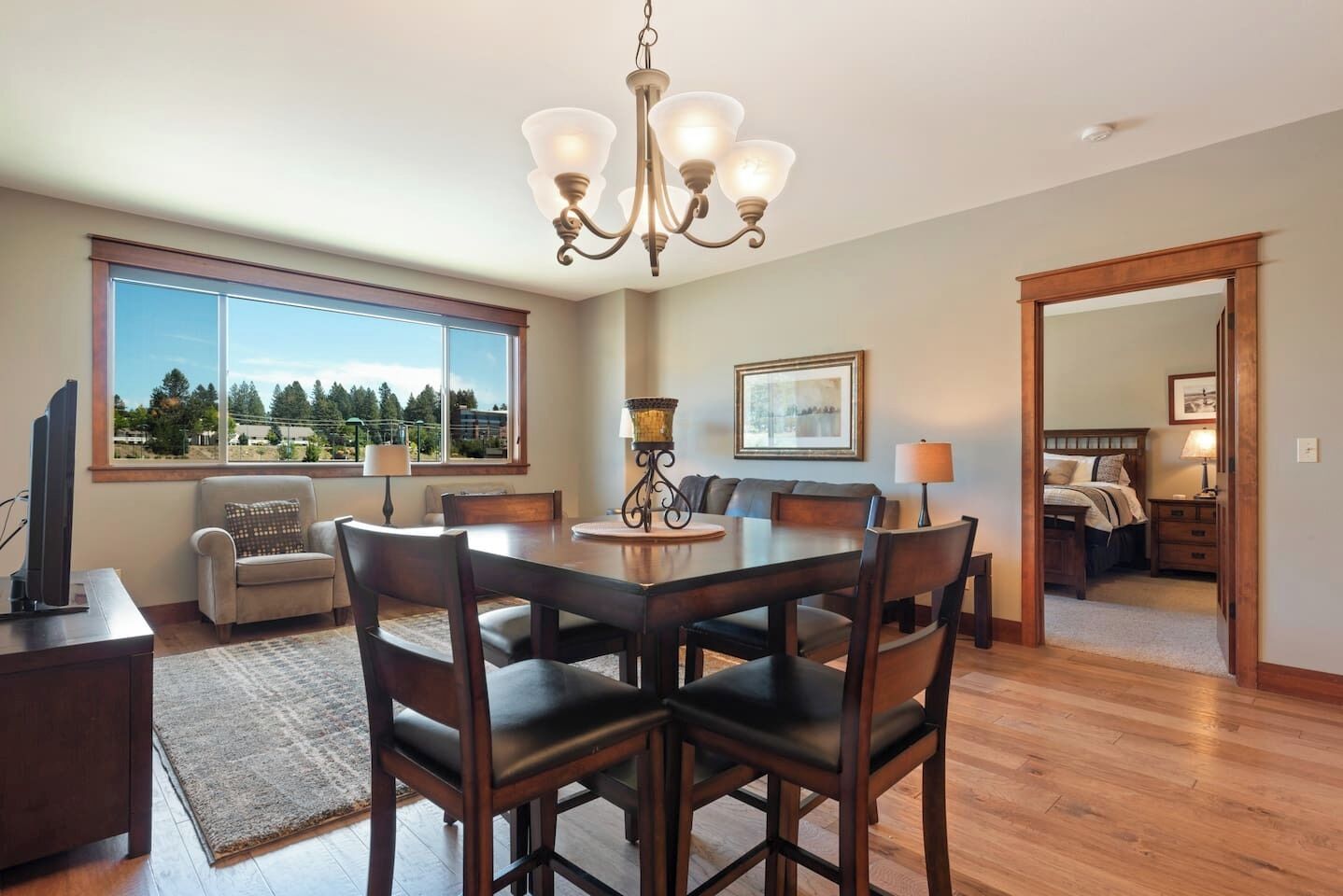 A spacious living room with a large window, a dining area with a chandelier, and a bedroom visible through an open doorway.
