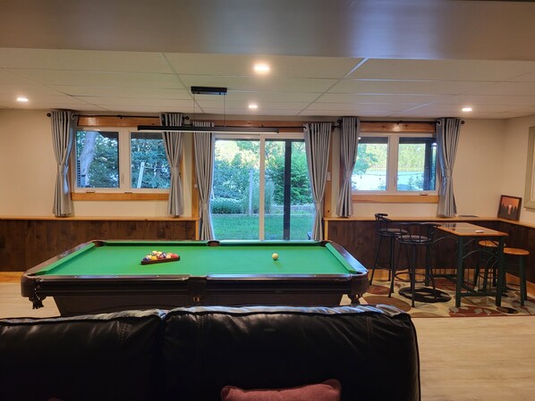 Une table de billard avec des billes, un canapé en cuir, une table à manger avec des chaises et des fenêtres avec des rideaux.