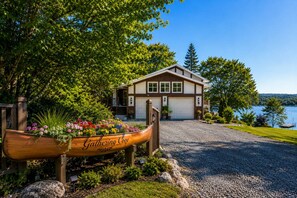 Une maison avec une allée en gravier, un panneau en bois en forme de bateau sur lequel on peut lire « Gathering Cove », et une vue sur le lac.
