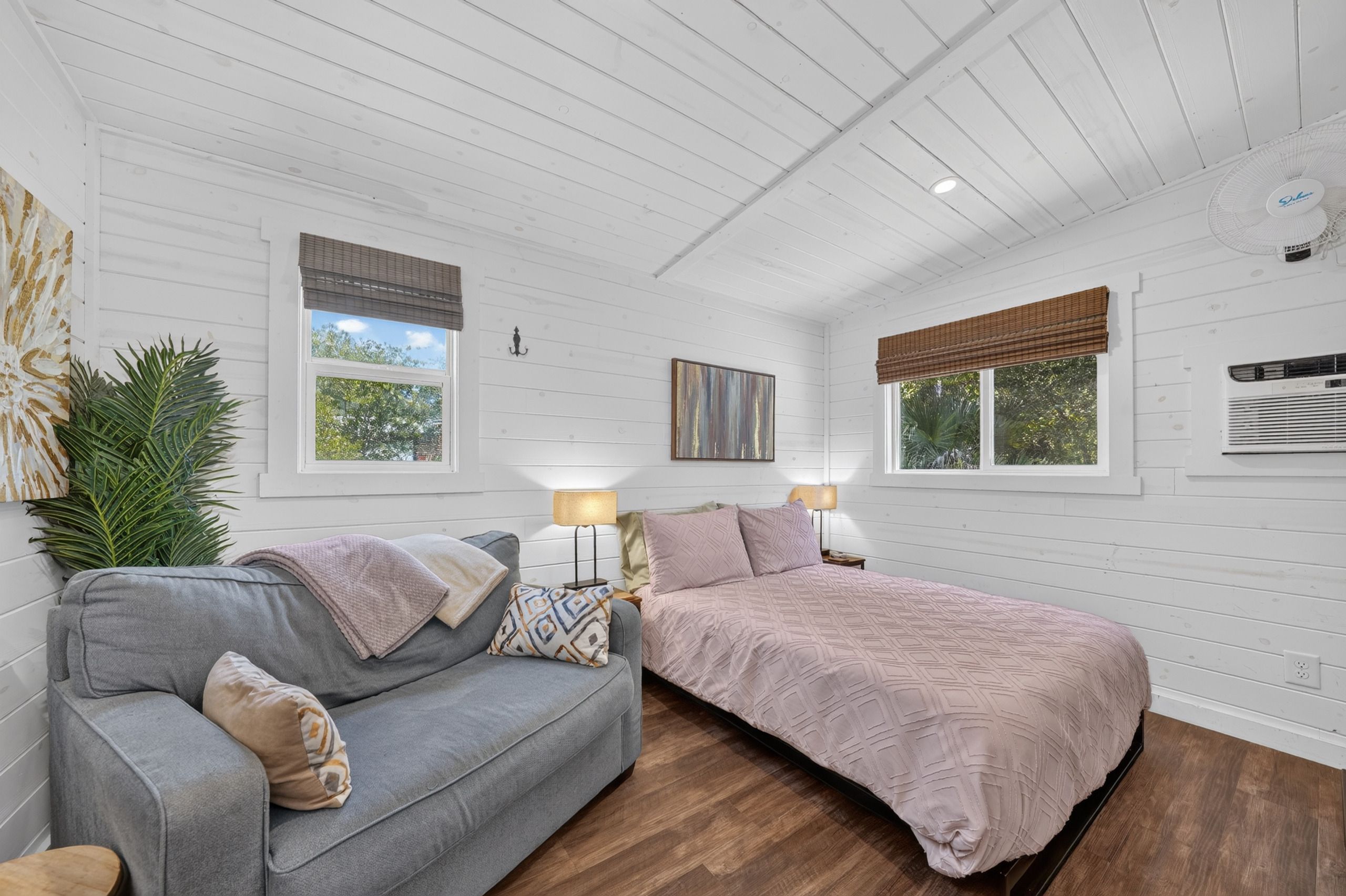 Ein Schlafzimmer mit grauem Sofa, einem Bett mit gemustertem Bettbezug, zwei Nachttischlampen, einem Bild an der Wand und einem großen Fenster mit Blick auf Bäume.