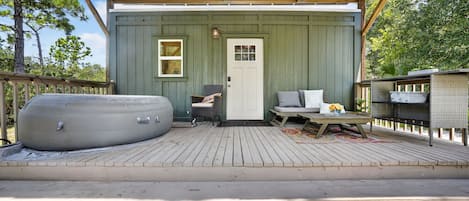 A covered outdoor seating area with a hot tub, a wooden deck, and a small building with a door and windows.