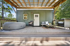 A covered outdoor seating area with a hot tub, a wooden deck, and a small building with a door and windows.