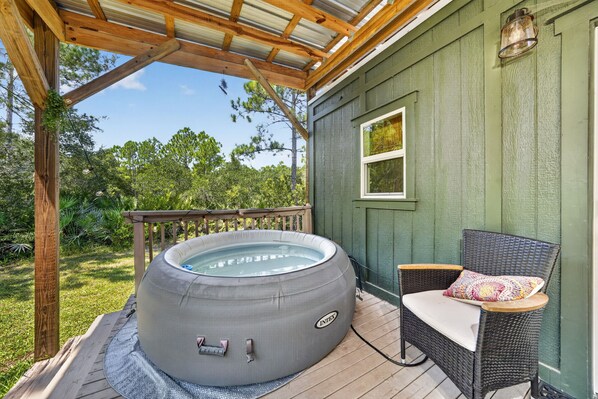 A hot tub on a wooden deck with a wicker chair and a small table, surrounded by greenery.