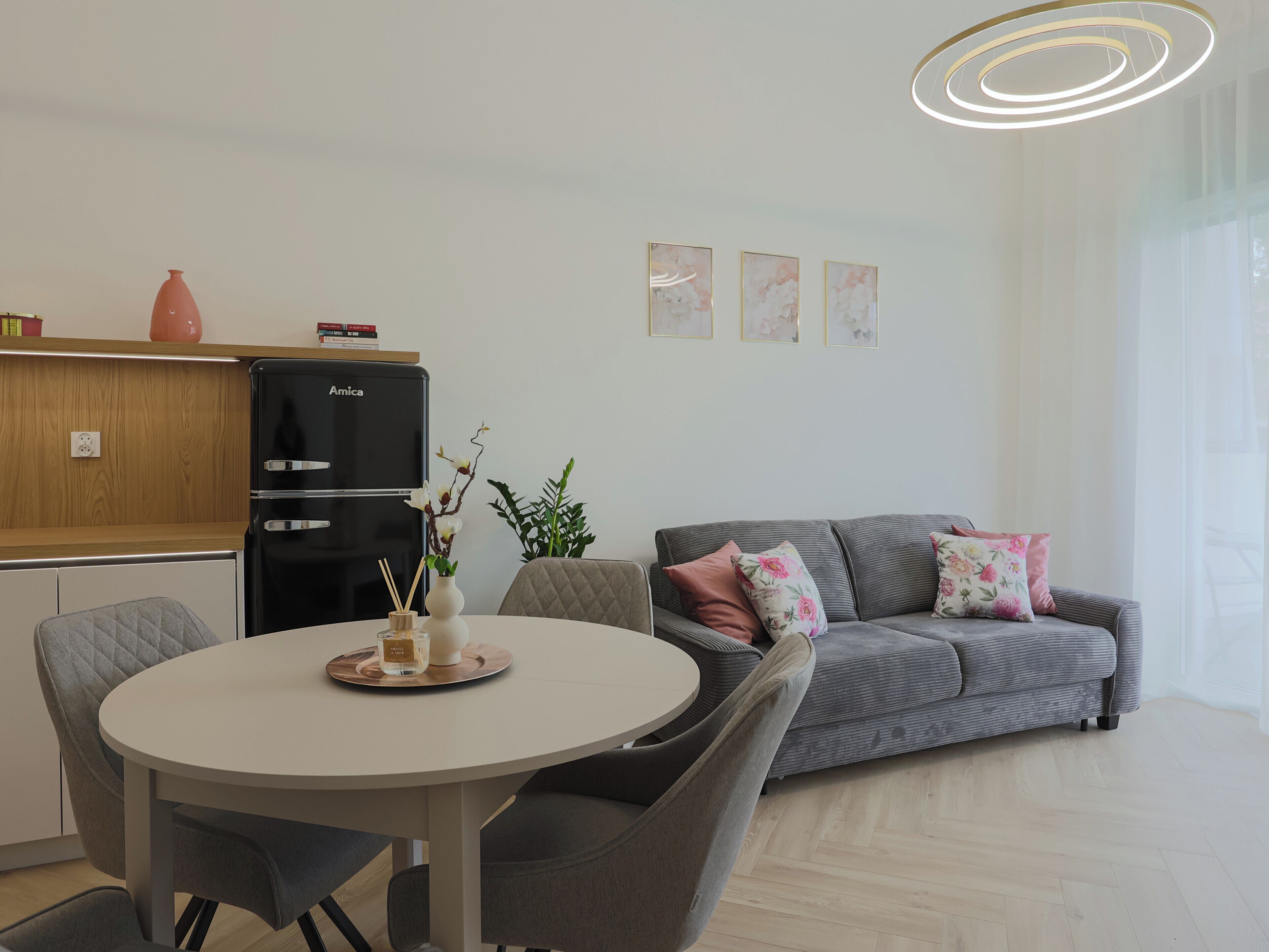 Un espace de vie moderne comprenant un coin repas, un canapé gris agrémenté de coussins à motifs floraux et un réfrigérateur noir.