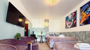 Habitación de hotel con televisor de pantalla plana, dos camas con cabeceras a juego, una mesita con planta y una zona de estar con sillones acolchados.