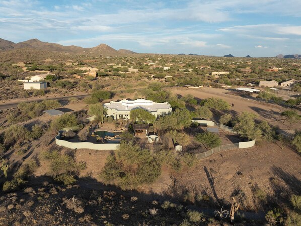 Una casa gran i moderna amb piscina, envoltada per un paisatge desèrtic amb vegetació escassa i muntanyes a la distància.