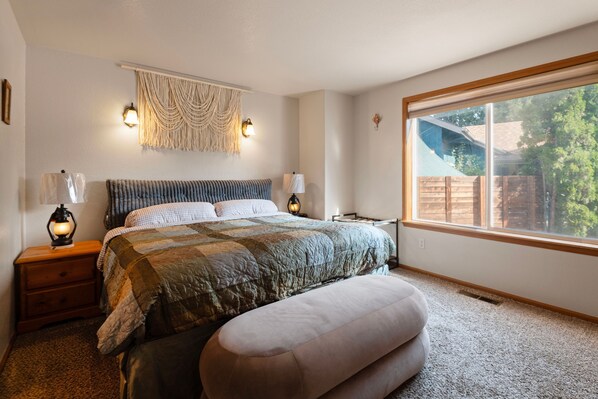 A bedroom with a large bed, bedside lamps, a wall-mounted macrame, and a window with a view of the outdoors. - 30+ Days: Quiet 3BR Home, Orchard District - Bend (Bend)