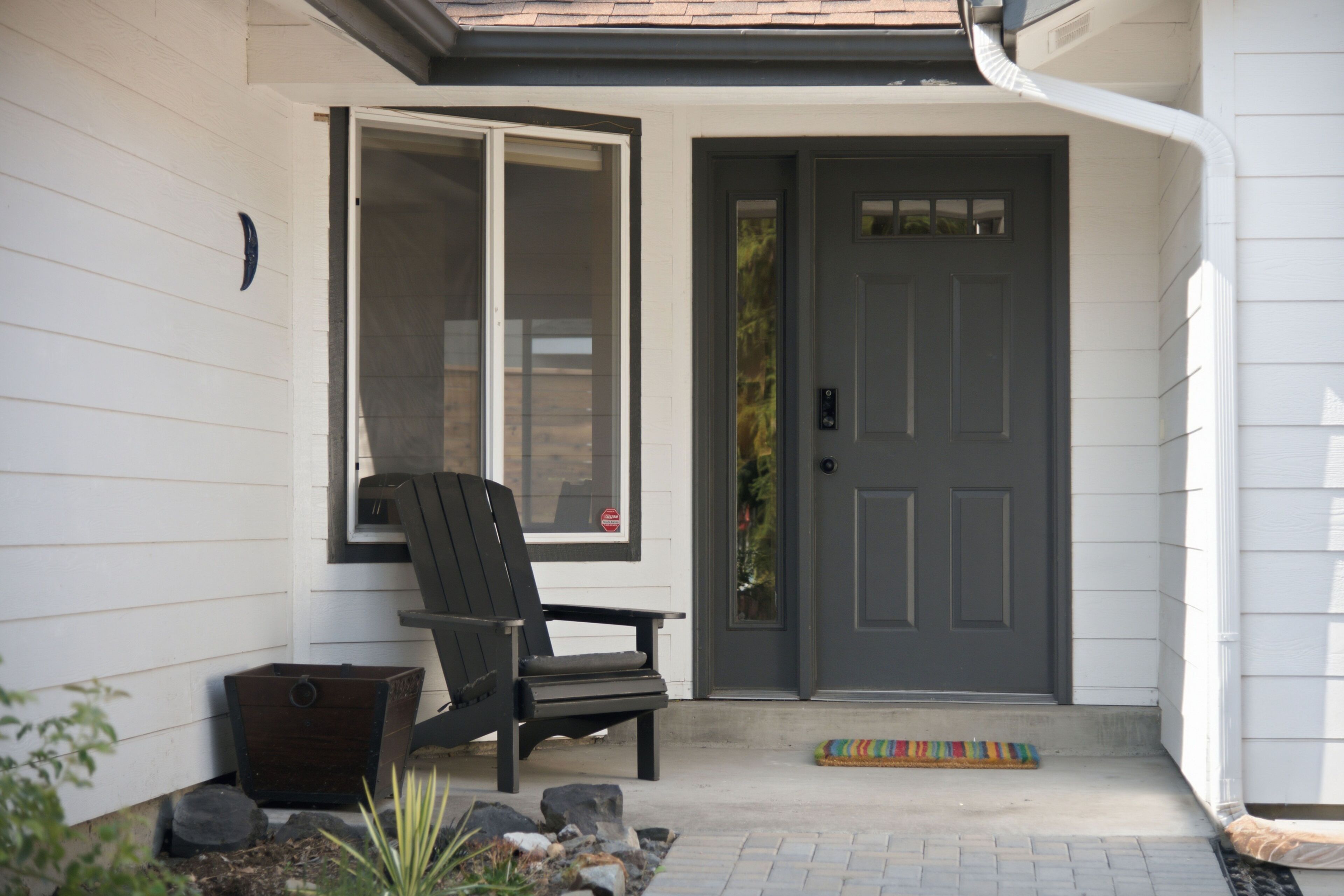 A front porch with a dark door, a sliding glass door, a wooden chair, a small wooden chest, and a colorful doormat.