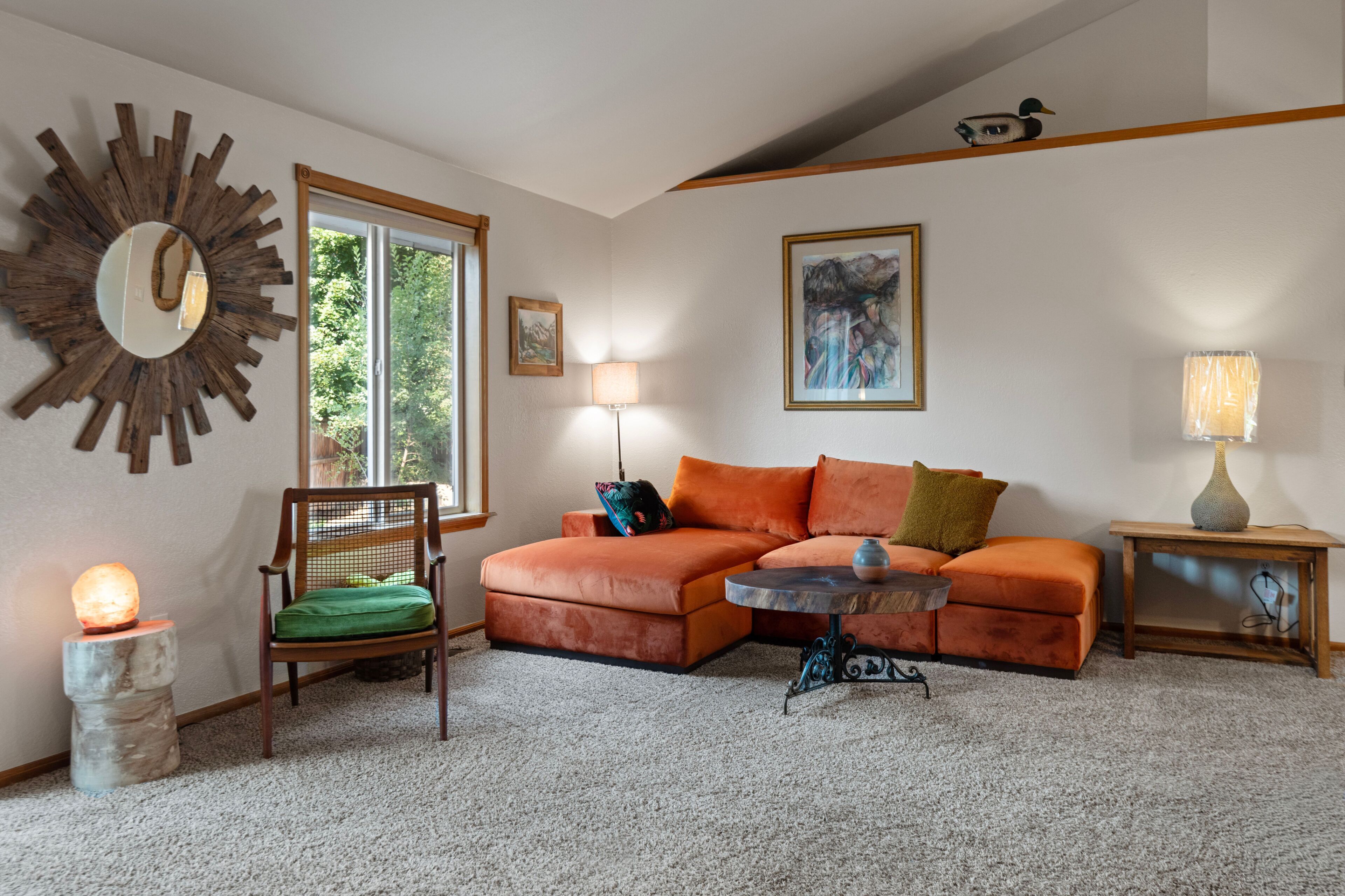 A living room with a sectional sofa, a round coffee table, a chair, a lamp, a painting, and a mirror on the wall.