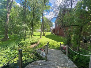 A wooden bridge leading to a grassy area with a red building in the background. - The Farm House at Mitchell Ponds (Knox)