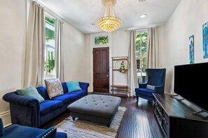 Uma sala de estar com sofá azul, otomano e poltrona, televisão e um lustre.