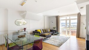 Un saló modern amb una taula de dinar de vidre, un sofà amb coixins, una finestra gran amb cortines i una vista a la ciutat.