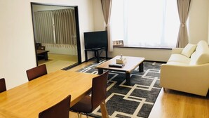 A modern hotel room with a dining table, chairs, a sofa, a TV, and a large window with curtains. - Minami Ward, Sapporo—10 or more people / Free shuttle service available / Perfect for groups and families with children! Free parking for 4 cars / We can arrange rental cars! (札幌市minami-ku,fujino)