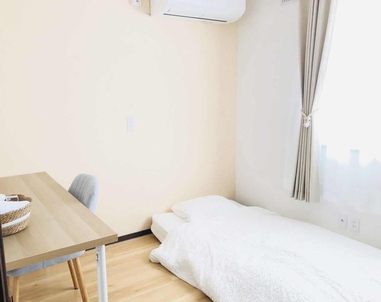 A small, single-room hotel room with a wooden table, a single bed, a chair, and a window with curtains.