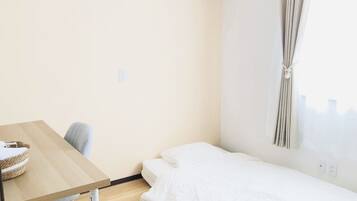 A small, single-room hotel room with a wooden table, a single bed, a chair, and a window with curtains.