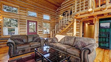 L’intérieur d’une cabane en rondins présente un salon avec deux canapés en cuir, une table basse en verre et un escalier.