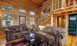 A log cabin interior with a living room featuring two leather sofas, a glass coffee table, and a staircase.