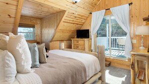 A wooden cabin bedroom with a bed, pillows, a TV, and a balcony with curtains. - Sunny Rise Cabin | Hot Tub, Pet Friendly, Fire Pit & Views! (Rutherfordton)