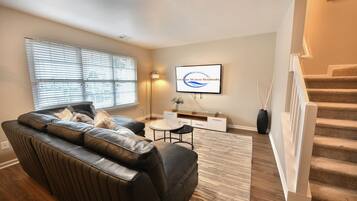 A living room with a leather sofa, a flat-screen TV, a small table, and a staircase.