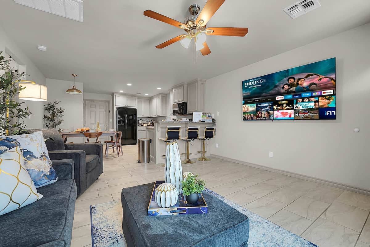 A modern living room with a sofa, a coffee table with decorative items, a ceiling fan, and a wall-mounted TV displaying various channels.