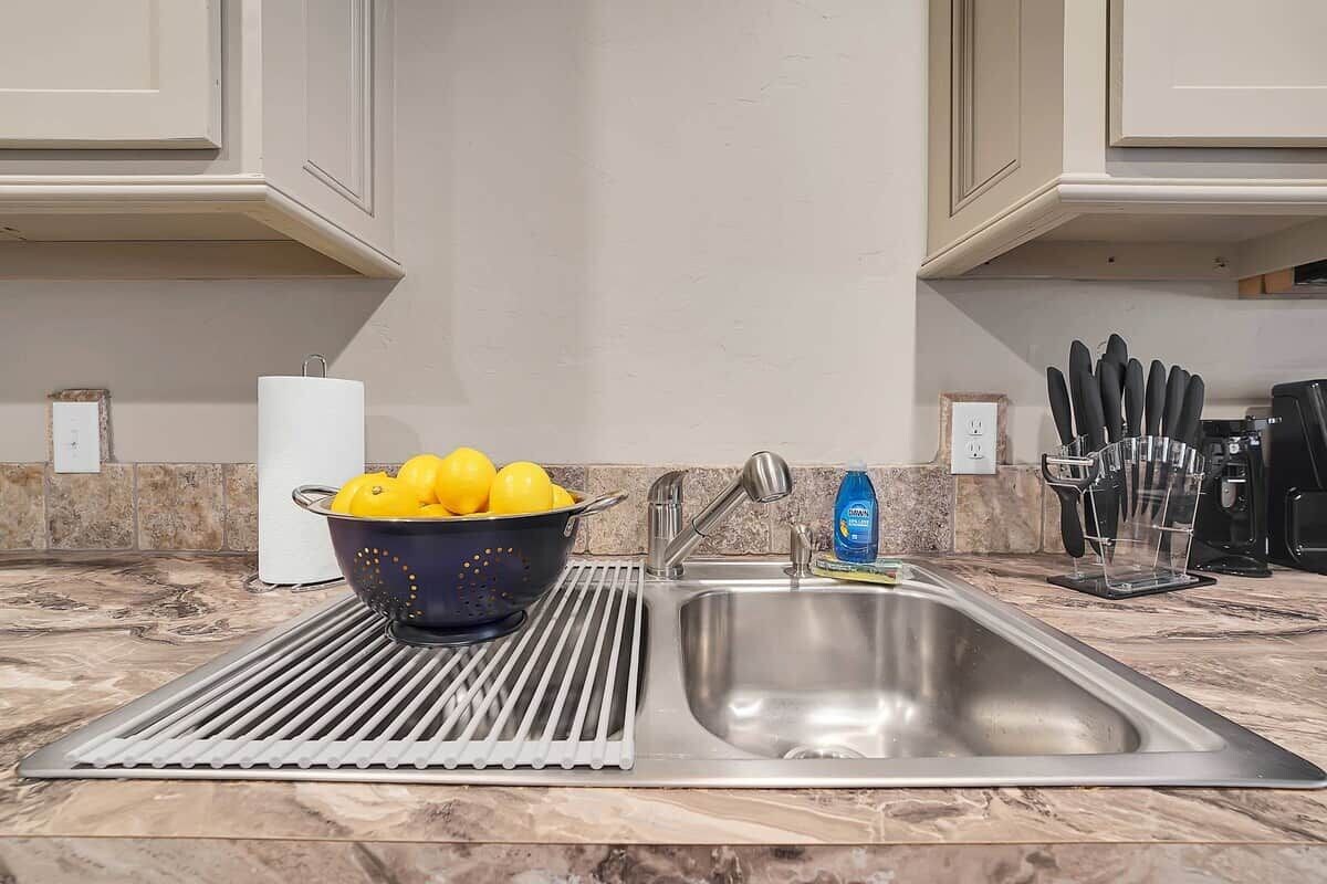 A kitchen with a stainless steel sink, a colander with lemons, a roll of paper towels, and a knife block.
