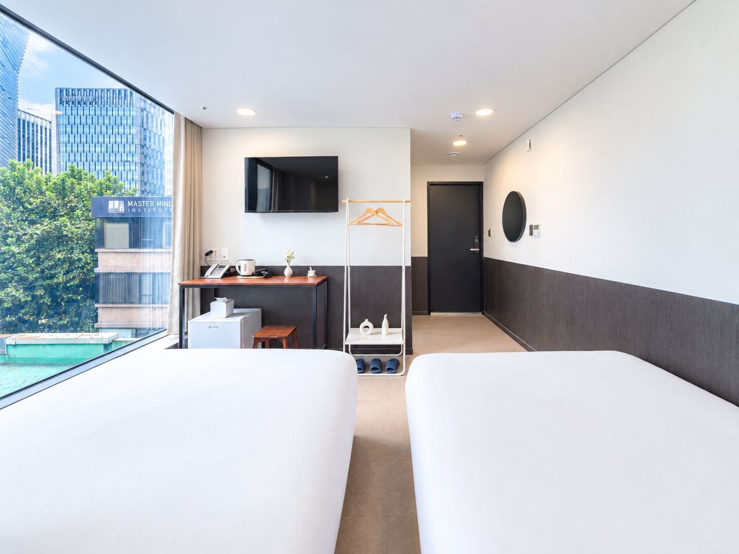 Habitación de hotel con dos camas, un escritorio con teléfono, televisor y vista a un edificio y árboles.