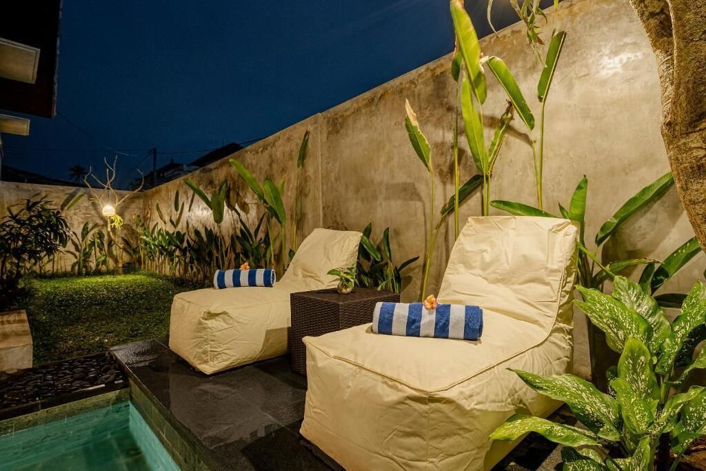 Un área junto a la piscina con dos sillas cubiertas, una pequeña mesa y diversas plantas.