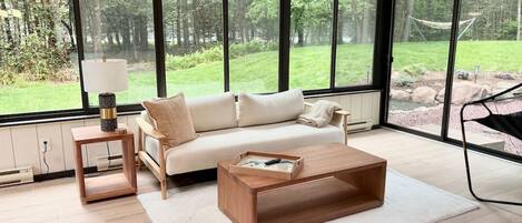 Un salón moderno con sofá, mesa de centro y mesita auxiliar, ventanas amplias con vista a un jardín.