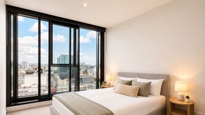 A bedroom with a large window, a bed with pillows, two bedside tables with lamps, and a view of the cityscape. - Skyline Sanctuary | Luxe Valley Apartment (Fortitude Valley)