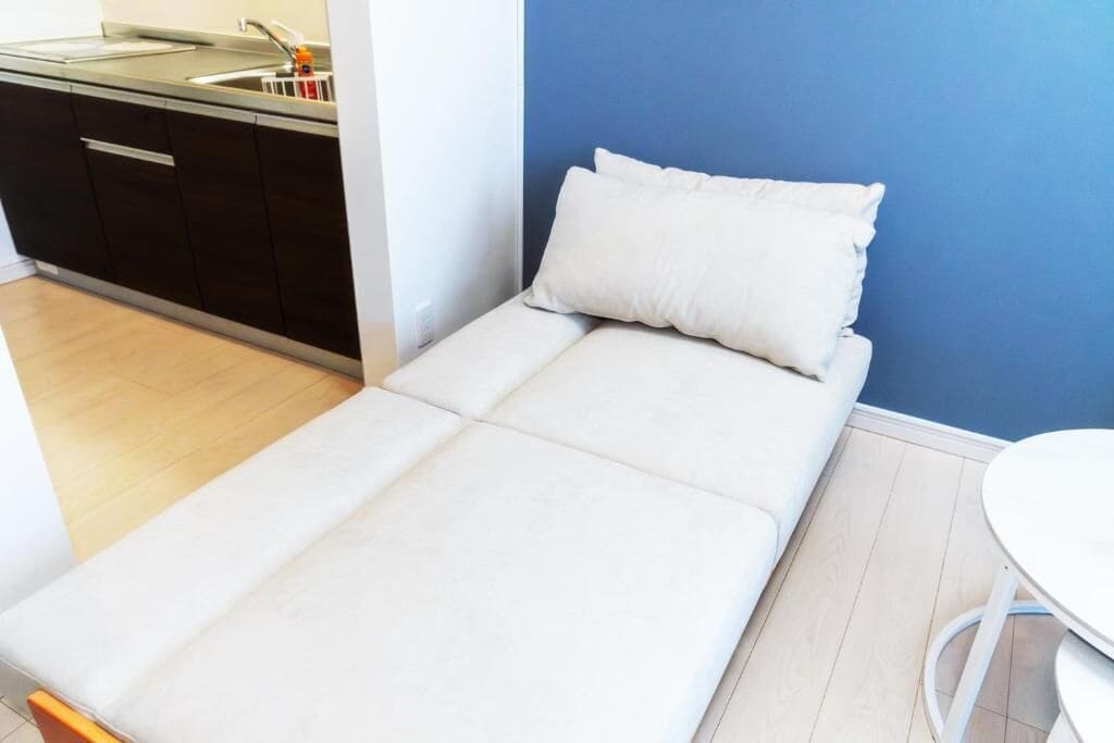 A small, single-bed room with a kitchenette, a white mattress, and a blue wall.