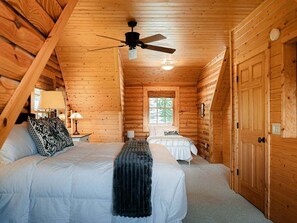 Quarto de cabana rústica com cama, uma cama menor, ventilador de teto, janela e paredes de madeira.