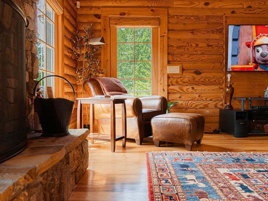 L’intérieur d’une cabane en rondins, avec une cheminée en pierre, des murs en bois, une grande fenêtre, un fauteuil en cuir, un petit pouf et un téléviseur.