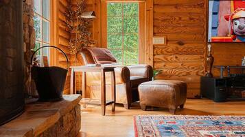 L’intérieur d’une cabane en rondins, avec une cheminée en pierre, des murs en bois, une grande fenêtre, un fauteuil en cuir, un petit pouf et un téléviseur.