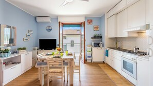Eine moderne Küche mit weißen Schränken, einem Essbereich mit Tisch und Stühlen, einem Fernseher und Blick durch eine Glastür nach draußen.