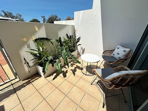 En balkon med krukker med planter, et lille rundt bord og to fletstole, samt en hvid væg.